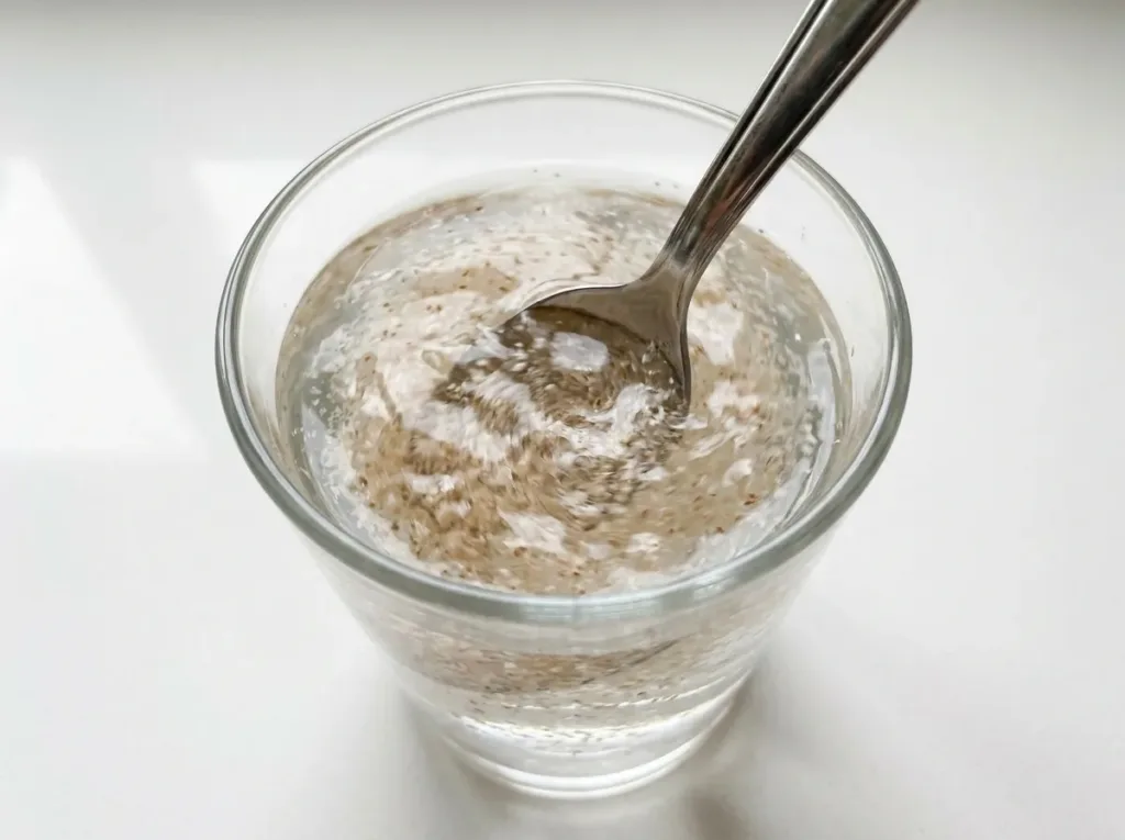 metal spoon stirring psyllium husk powder vigorously into water in a clear glass showing immediate stirring technique